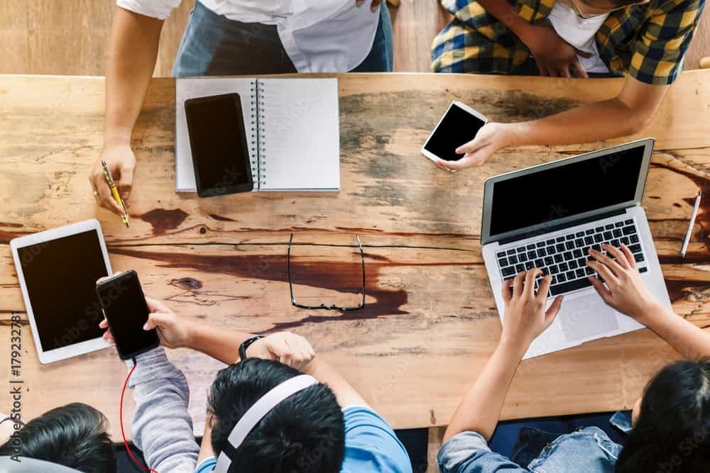 A group of people sitting around a table with laptops and cell phones, working on computer tech projects.