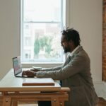 Man with glasses working on a laptop at a wooden desk by a brick wall and window.