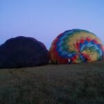 Two deflated hot air balloons lie on grassy field at dusk, with a dark balloon on the left and a colorful one on the right. A few people stand near the colorful balloon.