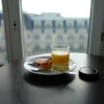 Glass of orange juice, a pastry, and a slice of meat on a plate with a spoon rest on a round table near a window with a city view.