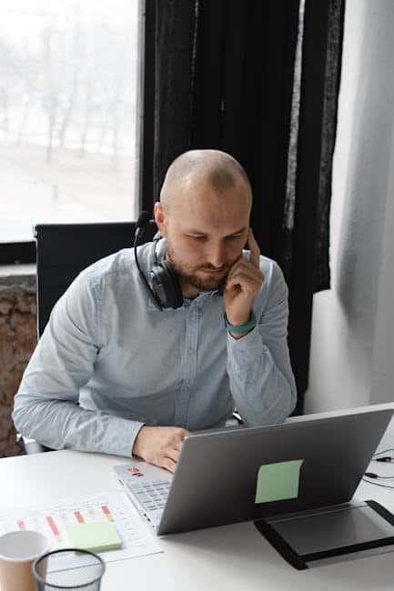 A man with headphones around his neck sits at a desk, looking at a laptop and touching his face, appearing deep in thought as he tackles IT support helpdesk tasks, with documents and a coffee cup nearby.