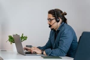 Man wearing a headset and glasses works on a laptop at a desk with a notebook, plant, and cup beside him in a modern office setting, providing IT Support Now to colleagues and clients.