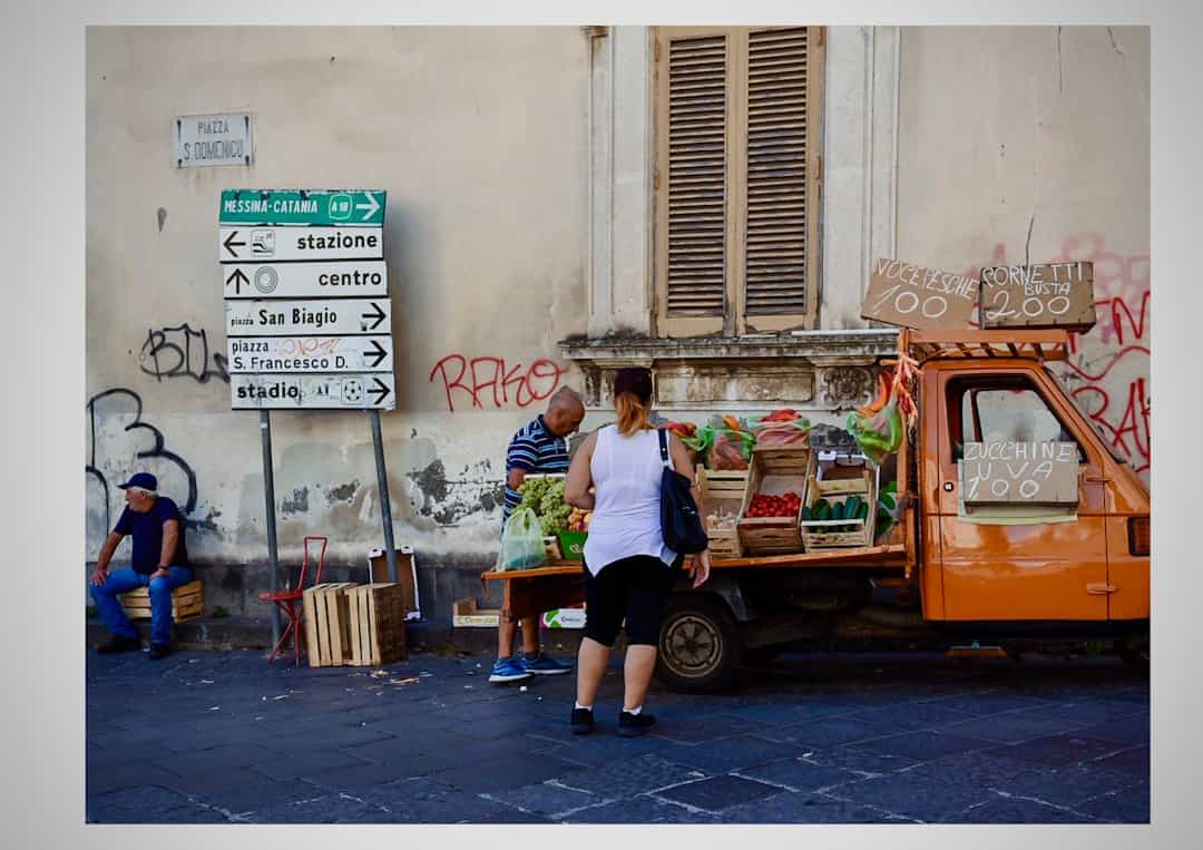 A woman buys produce from a vendor at an orange truck market stall on a city street; street signs and graffiti are visible on the wall behind them.